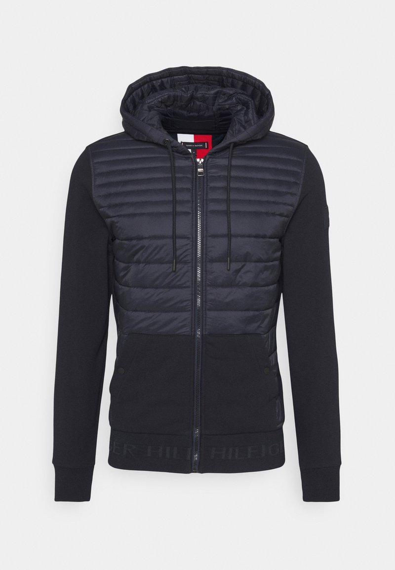 Navy hooded jacket with a quilted upper section, smooth lower section, front zipper, and logo detailing along the hem.