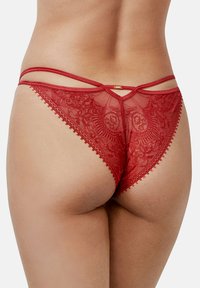 String en dentelle rouge avec tissu semi-transparent, présentant un motif floral, doublés de fines bretelles spaghetti et des bords festonnés le long de la ceinture.