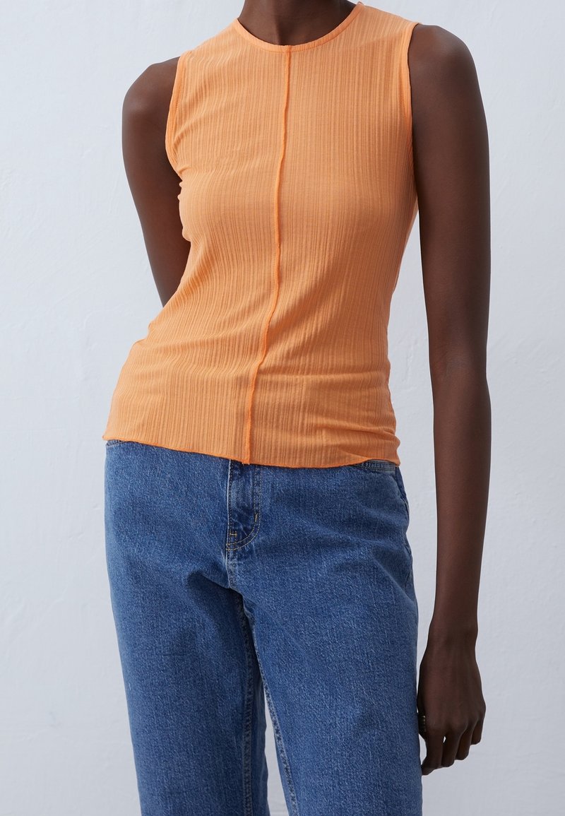 Ribbat, ärmlös orange topp med hög hals och mittsöm, kombinerad med blå jeans. Tyget har synlig struktur och en åtsittande form.