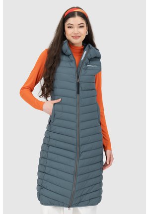 Frau trägt lange graue gesteppte Weste mit Frontreißverschluss und hohem Kragen über orangefarbenem Langarmshirt, posiert mit einer Hand in der Tasche.