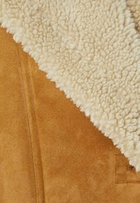 Hnědý semišový materiál s krémovým, nadýchaným šálovým límcem. Textura kontrastuje mezi hladkým a měkkým. Obsahuje detail knoflíku a kapsy.