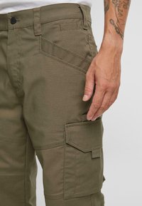 Pantalones cargo verde oliva con un tejido texturizado, bolsillos laterales y una silueta ajustada. Costuras visibles y un detalle en la cinturilla.