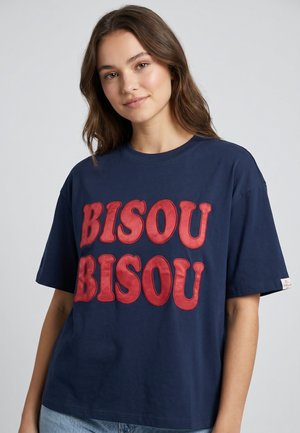 Woman with long brown hair wearing a navy blue T-shirt with large red text "BISOU BISOU" and light blue jeans against a plain background.