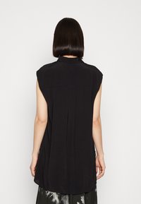 Person med kort, rakt hår som bär en ärmlös svart topp, sedd bakifrån mot en plain ljus bakgrund.