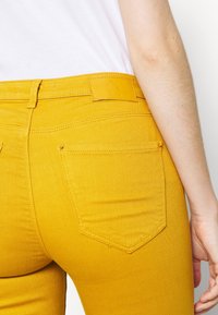 Keltainen farkkuhousut, joissa on slim fit -leikkaus. Vyötä ympäröi nahkapaikka ja takana on kaksi taskua, joissa on ainutlaatuinen, terävä muoto.