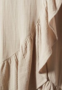 Tissu beige à texture douce, avec des coutures diagonales et des volants délicatement froncés créant des plis superposés.