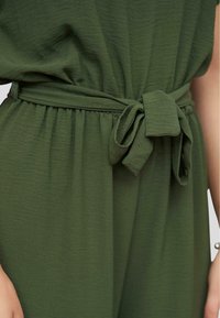 Green fabric dress gathered at the waist with a tied self-fabric belt, showing part of a person's arm with a pearl bracelet.