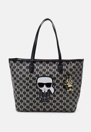 Czarna torba typu tote z powtarzającym się białym geometrycznym wzorem, z postacią z kreskówki w okularach przeciwsłonecznych oraz dopasowanym breloczkiem jako akcesorium.