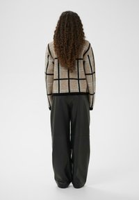 Bēša adīta džemperis ar melnu un gaiši zilu režģa rakstu, kombinēts ar brīvi pieguļošām melnām ādas biksēm. Skats no aizmugures, modele ar garām cirtainām matiem.