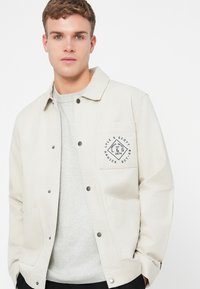 Giacca beige chiaro con chiusura a bottoni, due tasche laterali e una tasca sul petto con una toppa del logo nera. Indossata sopra una maglietta grigia.