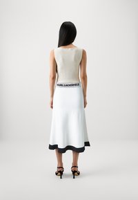 Strikket ermeles topp i lys beige, kombinert med en hvit skjørt med svart kant. Svart logo midjebånd, slank silhuett og glatt tekstur.