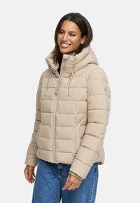 Beige pufferjacka med huva, quiltad textur, främre dragkedja och justerbara snoddar. Matchad med blå jeans för en avslappnad look.
