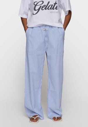 Persona che indossa pantaloni larghi a righe verticali blu e bianche con coulisse, sandali bianchi e una maglietta bianca con la scritta "Gelati".
