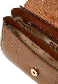 Intérieur de sac à main en similicuir marron texturé avec ferrures dorées, comprenant deux compartiments et le nom de la marque "GUESS" visible sur la doublure.