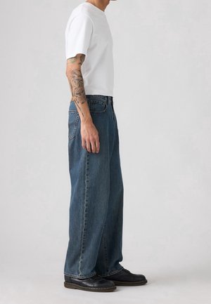 Mężczyzna w białej koszulce, luźnych niebieskich dżinsach i czarnych butach, stojący bokiem, z widocznym tatuażem na prawej ręce na jednolitym tle.