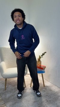 Marineblå hettegenser med rosa logo, svarte jeans og svart-hvite joggesko. Står på et beige teppe, med en hvit stol og bøker bak.