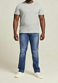 Homme portant un t-shirt gris clair à manches courtes, un jean bleu et des chaussures blanches, debout avec les bras détendus le long du corps.