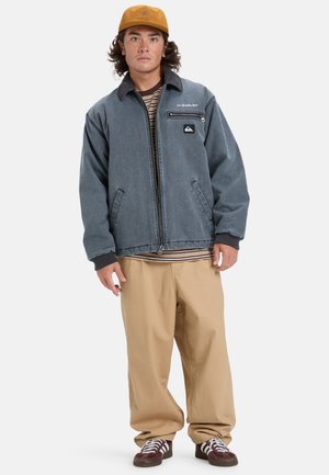 Veste en denim bleue avec zip sur le devant, deux poches latérales et un patch logo. Associée à un pantalon beige et des chaussures brunes. Chemise rayée visible en dessous.