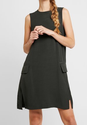 Mujer con el pelo largo y trenzado, vestida con un vestido negro sin mangas con bolsillos laterales y aberturas, de pie con las manos entrelazadas delante.