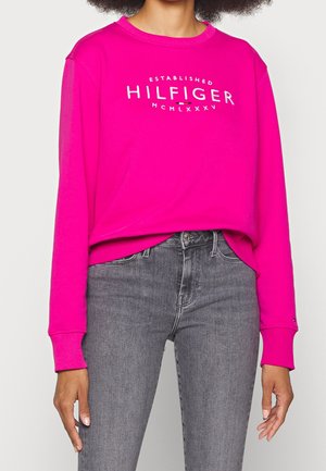 Woman wearing bright pink long-sleeve shirt with "Hilfiger" text and gray jeans, shown from neck to hips against plain background.