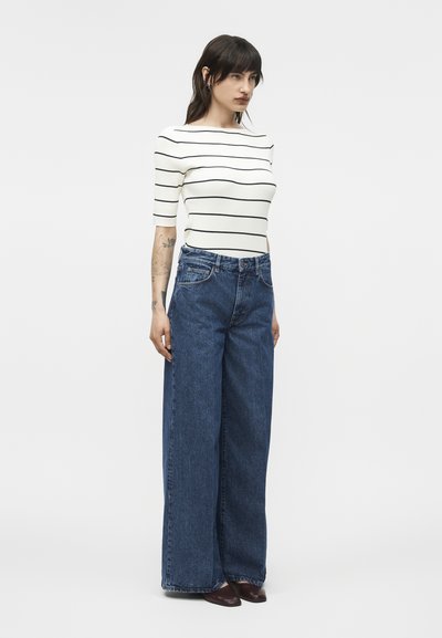 Žena stojaca, oblečená v bielom tričku s krátkym rukávom a čiernymi horizontálnymi pruhmi, modrých džínsach s vysokým pásom a širokými nohavicami a tmavo hnedých topánkach.