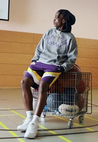 Sudadera gris con diseño de zigzag morado, pantalones cortos morados y amarillos, y zapatillas blancas. Sentado en un carrito de metal lleno de pelotas deportivas.