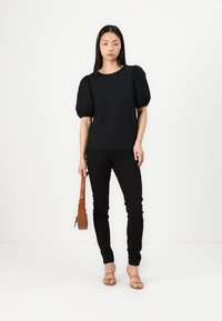 Svart puffermed topp med rund hals, kombinert med smale svarte jeans og beige stroppsandaler, holder en brun skulderveske.