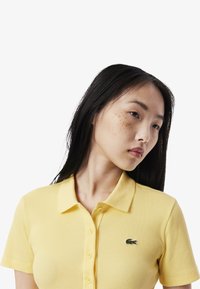 Gul randig polo-shirt med krage, med en liten broderad grön logotyp på vänster bröst. Korta ärmar.