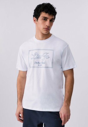 Giovane uomo con capelli ricci scuri che indossa una maglietta bianca "Liu Jo Swim Club" e pantaloni scuri davanti a uno sfondo chiaro uniforme.