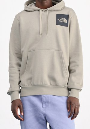 Osoba nosząca jasnoszarą bluzę z kapturem The North Face z przednią kieszenią i ściągaczami, w zestawieniu z jasnoniebieskimi spodniami, na jednolitym białym tle.
