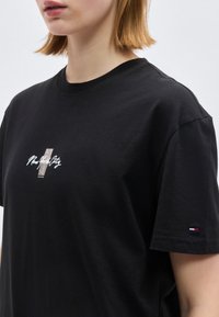 Άτομο που φοράει μαύρο t-shirt με μικρό κείμενο "New York City" και λογότυπο στο στήθος και μικρό έμβλημα σημαίας στο μανίκι.