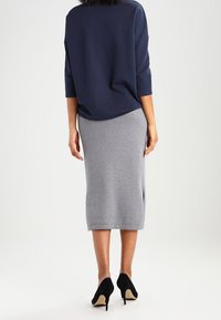 Mujer vestida con una blusa azul marino, una falda gris hasta la rodilla y unos zapatos de tacón negros, de pie de espaldas contra un fondo blanco.