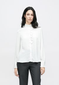 Balta blūze ar garām piedurknēm, volānu apkakli un pogām priekšpusē, izgatavota no viegla auduma, kombinēta ar tumšām biksēm.