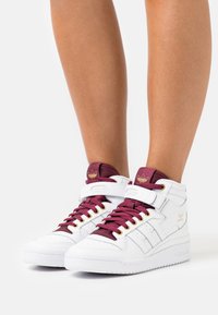 Höghöga vita sneakers med burgundy detaljer, lädermaterial, guldfärgade beslag och en vadderad krage med en logotyp på tungan.