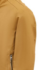 Mosterdgele jas van gladde, gestructureerde stof, met een ritssluiting pocket aan de linkerkant en nette stiksels langs de naden.