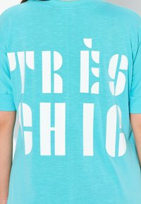 Tyrkysové tričko s výraznými bílými blokovými písmeny s nápisem "TRÈS CHIC" na zadní straně. Měkká, lehká tkanina s volným střihem.