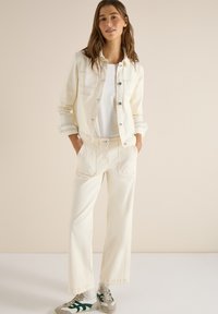 Jeune femme debout, les mains dans les poches, portant une veste en denim blanc cassé, un pantalon large assorti, un haut blanc et des baskets.