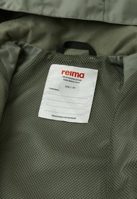 La giacca impermeabile verde presenta un interno con fodera in rete e un'etichetta con il marchio "reima" e le informazioni sulle taglie, inclusa la scritta "Io ero qui".