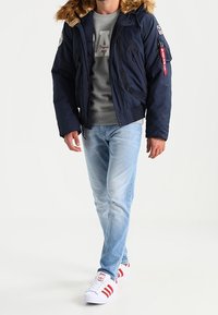 Hombre caminando con una chaqueta de piel sintética color marino con capucha, sudadera gris, jeans azul claro y zapatillas blancas con rayas rojas.
