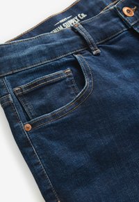 Jeans din denim albastru închis cu cusături vizibile. Au un buzunar mic în față, un buton din cupru și o etichetă a mărcii pe talie.