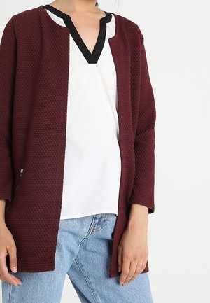 Giacca bordeaux con texture e colletto nero, indossata sopra una maglietta bianca, abbinata a jeans in denim azzurro chiaro. Maniche lunghe.
