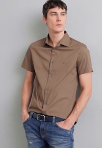 Cămașă maro cu mânecă scurtă și butoane, cu un logo subtil, asociată cu blugi denim albaștri și o curea neagră, pe un model pe un fundal gri.