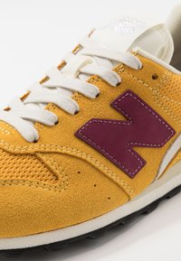 Gul mockasneaker med vita snören och en strukturerad meshtavla. Har en framträdande lila "N"-logotyp och en svart gummisula.