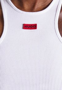 Närbild av en vit ribbad linne med en röd rektangulär lapp med texten "HUGO" på bröstet, bärs av en person med mörk hud.