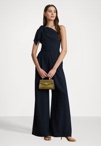 Mono azul marino con un diseño de un solo hombro y detalle de lazo. La modelo sostiene un pequeño bolso dorado. Textura suave, silueta de pierna ancha, tacones con tiras.