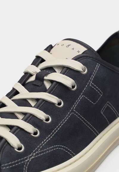 Pantof sport din piele intoarsa albastru marin cu cusaturi albe, șireturi albe, orificii metalice și o etichetă bej de brand pe limba.