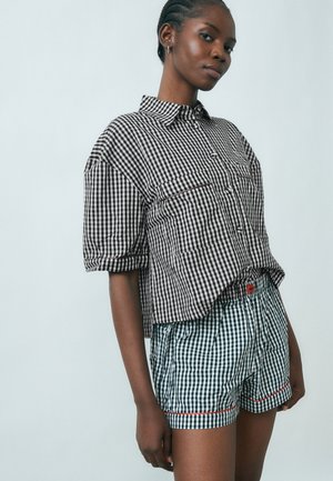 Femme portant une chemise à carreaux noir et blanc à boutons avec manches retroussées et un short assorti avec bordure rouge, se tenant devant un fond uni.