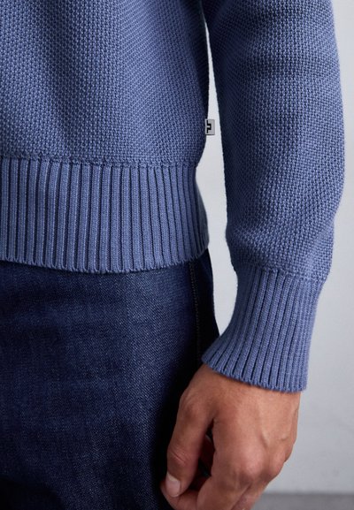 Strikket sweater i lys blå med et struktureret mønster. Har ribbede manchetter og kant. Bæres med mørke denimbukser.