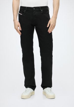 Uomo che indossa jeans neri a gamba dritta e sneakers bianche, in piedi con le mani rilassate lungo i fianchi davanti a uno sfondo semplice.