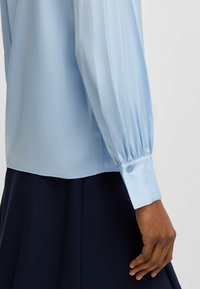 Blusa azzurro chiaro con maniche lunghe e gonfie e polsini con bottoni. Tessuto liscio e lucente. Abbinata a una gonna scura, che mostra una silhouette pulita.